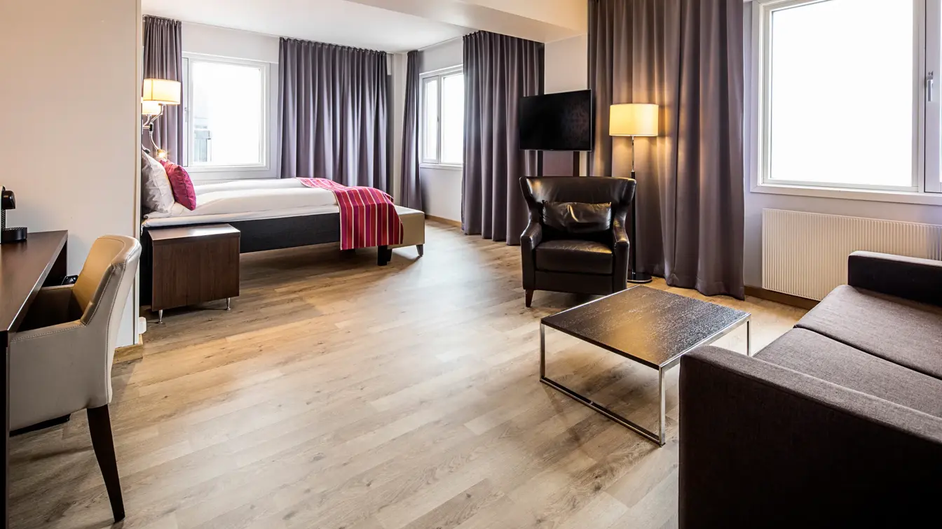 Brun sofa, lenestol, salongbord, vindu, grå gardiner, skrivebord, stol, dobbeltseng, nattbord.