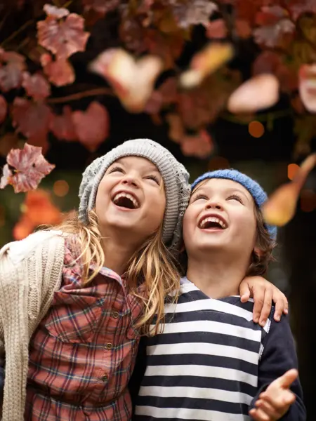 Shot of two cute kids laughing joyfully while watching autumn leaves falling from the trees
