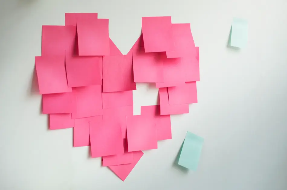 Blank pink sticky notes sticked in a heart shape on wall.