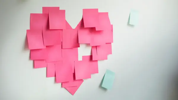 Blank pink sticky notes sticked in a heart shape on wall.