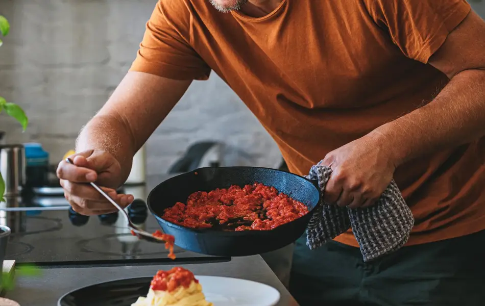 Handsome young happy Caucasian man making classic tagliatelle by putting sauce from a pan to pasta on a plate in a kitchen.