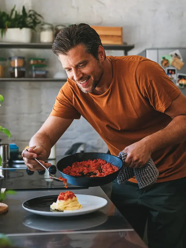 Handsome young happy Caucasian man making classic tagliatelle by putting sauce from a pan to pasta on a plate in a kitchen.