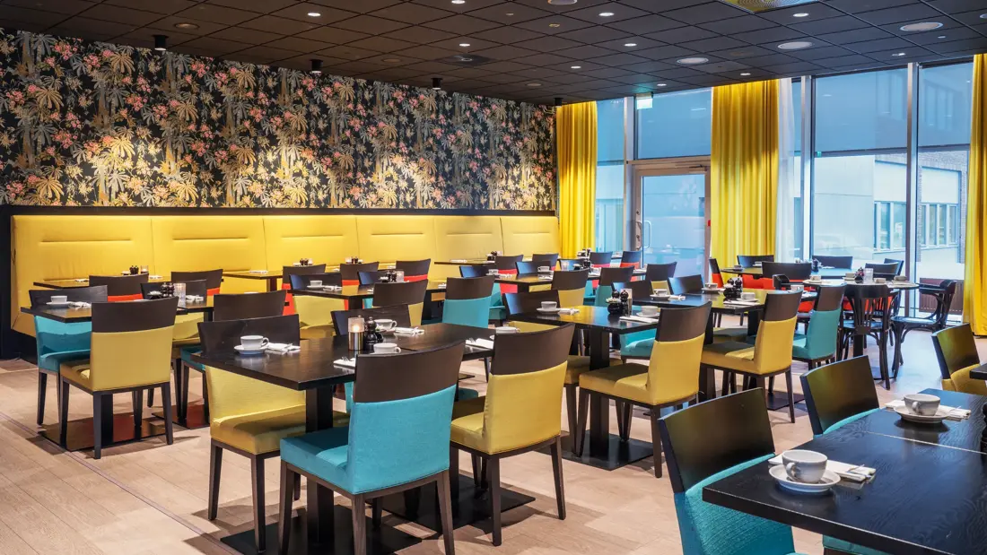 Tapetvegg med mønster, vindu fra gulv til tak, gule gardiner, spisebord med stoler, lys parkett. 