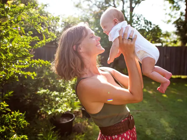 Kvinne smiler og løfter opp baby i hage på sommeren