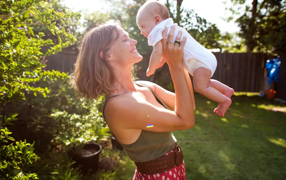 Kvinne smiler og løfter opp baby i hage på sommeren