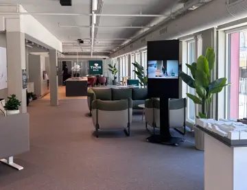 Lyst og moderne visningssenter for boligprosjektet Dronningkvartalet fra Thon Bolig med sittegrupper, store vindusflater, grønne planter, digital informasjonsskjerm og boligmodell utstilt i forgrunnen.