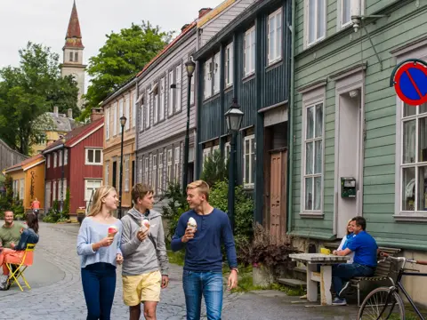 Tre ungdommer går og prater sammen og spiser is på Bakklandet i Trondheim. I bakgrunnen ses bygninger i ulike farger og folk som sitter på uteserveringer.