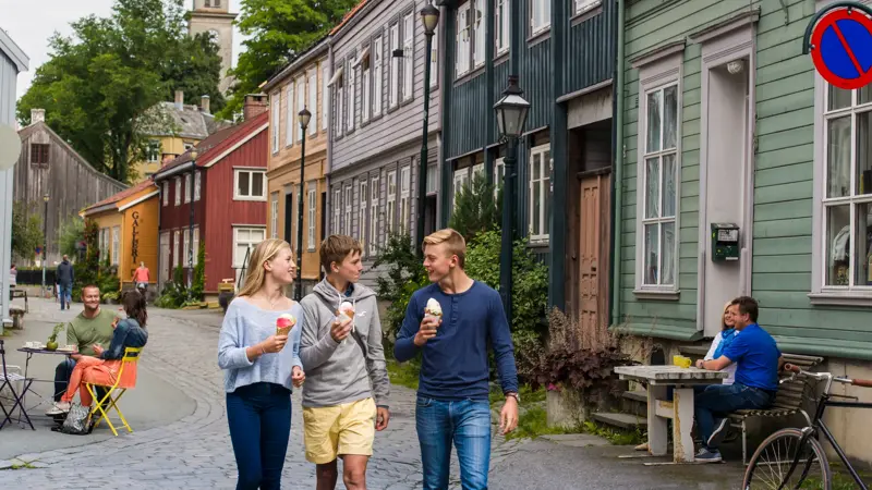 Tre ungdommer går og prater sammen og spiser is på Bakklandet i Trondheim. I bakgrunnen ses bygninger i ulike farger og folk som sitter på uteserveringer.