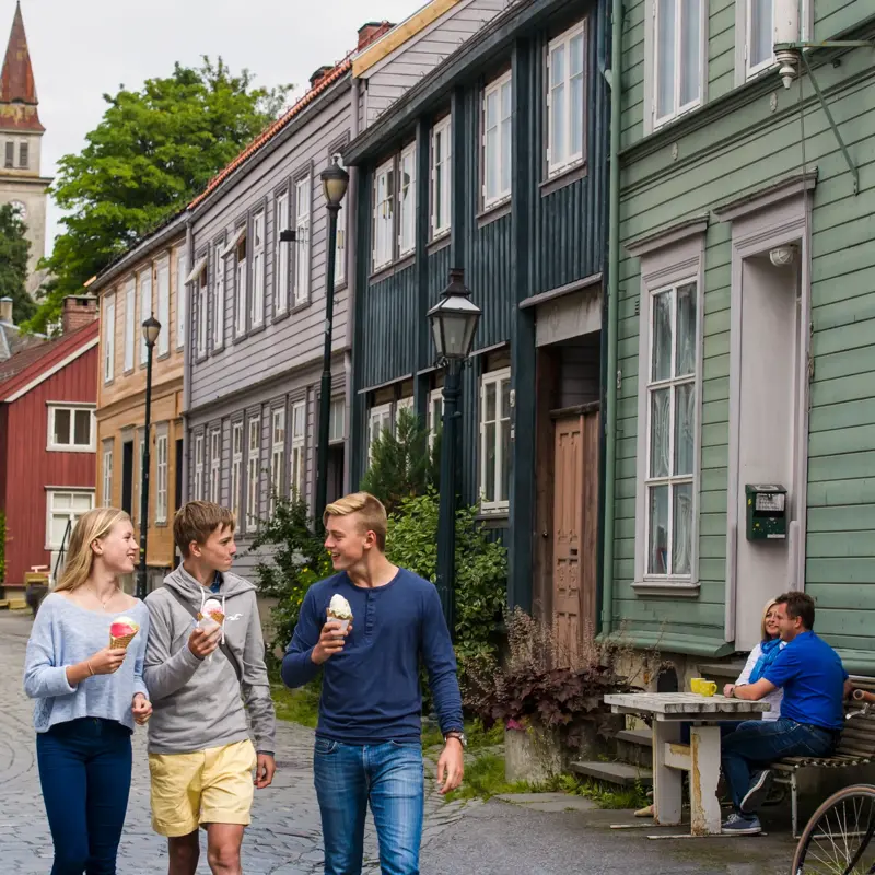 Tre ungdommer går og prater sammen og spiser is på Bakklandet i Trondheim. I bakgrunnen ses bygninger i ulike farger og folk som sitter på uteserveringer.