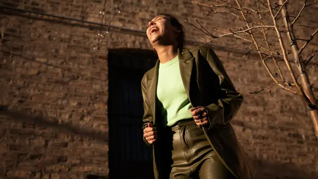 Dame med grønn jakke og bukse og lys grønn topp ler og ser opp mot himmelen utendørs