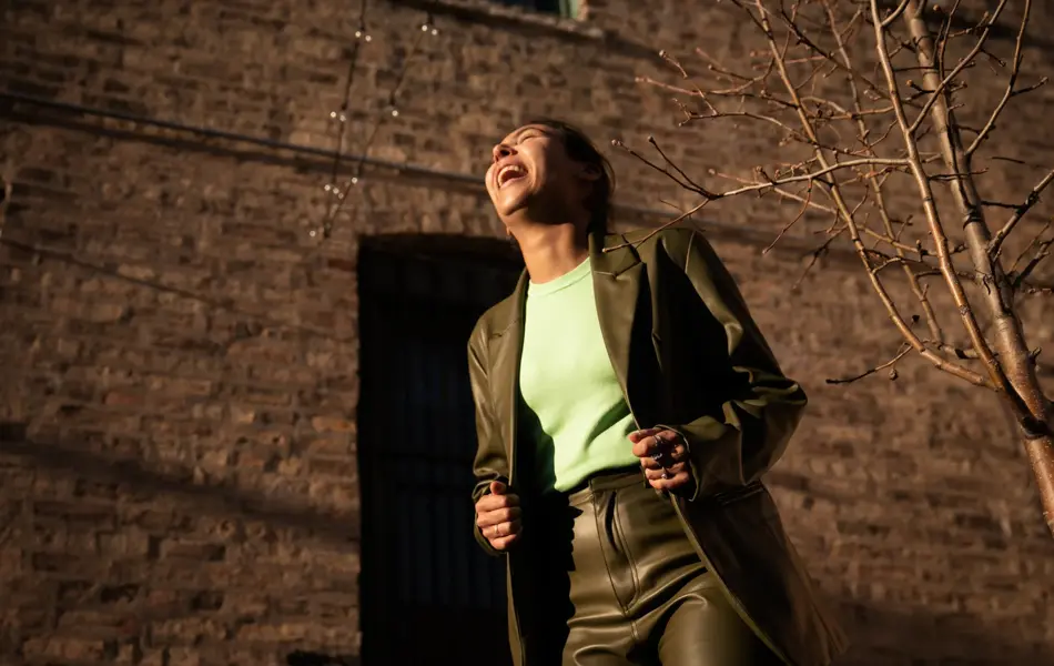 Dame med grønn jakke og bukse og lys grønn topp ler og ser opp mot himmelen utendørs