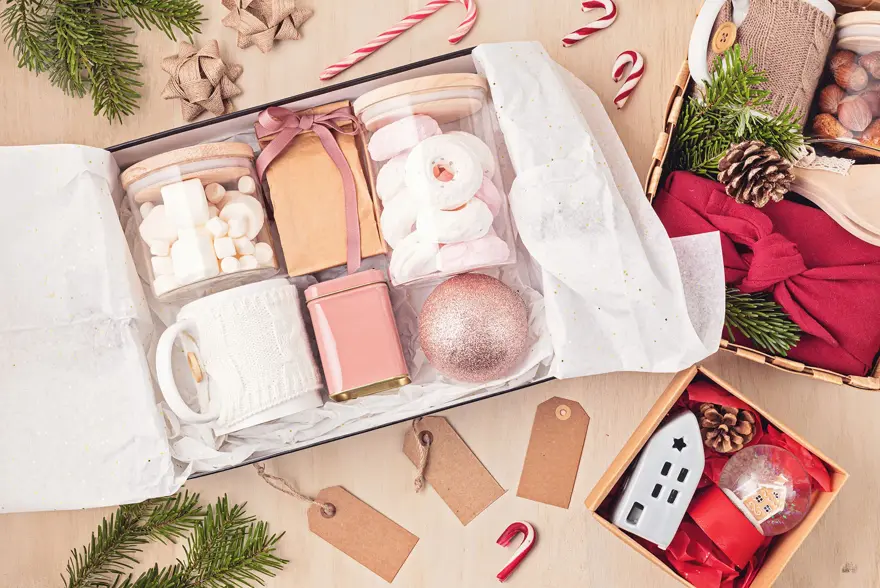 Preparing care package, seasonal gift box with marshmallow, tea,coffee or cacao box and christmas ornament. Personalized eco friendly basket for winter holidays. Top view, flat lay