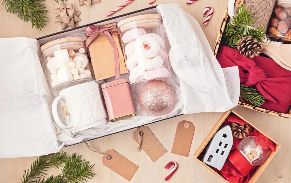 Preparing care package, seasonal gift box with marshmallow, tea,coffee or cacao box and christmas ornament. Personalized eco friendly basket for winter holidays. Top view, flat lay