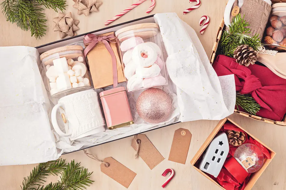 Preparing care package, seasonal gift box with marshmallow, tea,coffee or cacao box and christmas ornament. Personalized eco friendly basket for winter holidays. Top view, flat lay