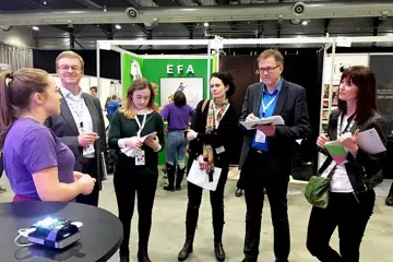 Fem medlemmer av en jury står i messehallen til Ungt Entreprenørskap og får presentasjon fra en ung gründer.