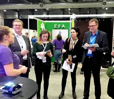 Fem medlemmer av en jury står i messehallen til Ungt Entreprenørskap og får presentasjon fra en ung gründer.