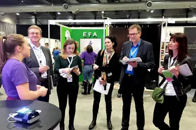 Fem medlemmer av en jury står i messehallen til Ungt Entreprenørskap og får presentasjon fra en ung gründer.