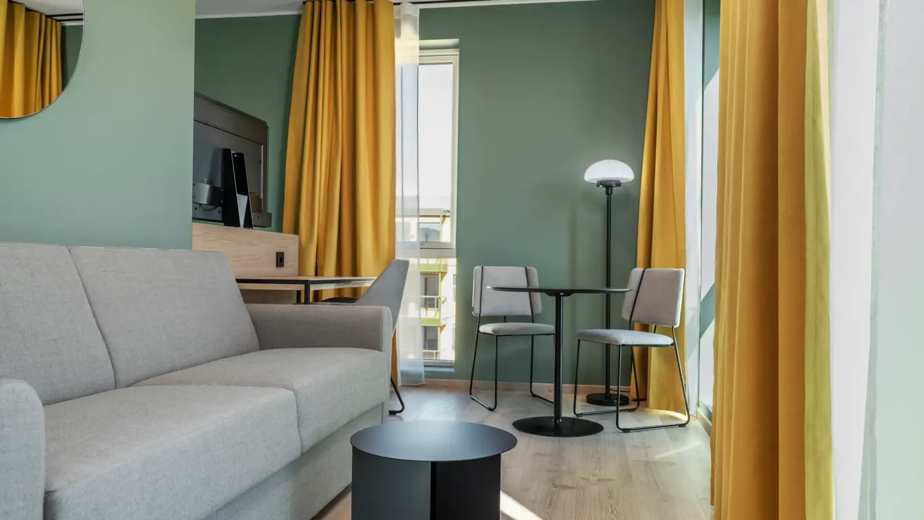 Standard queen rom med grå sofa, spiseplass, vinduer, gule gardiner og grønne vegger.