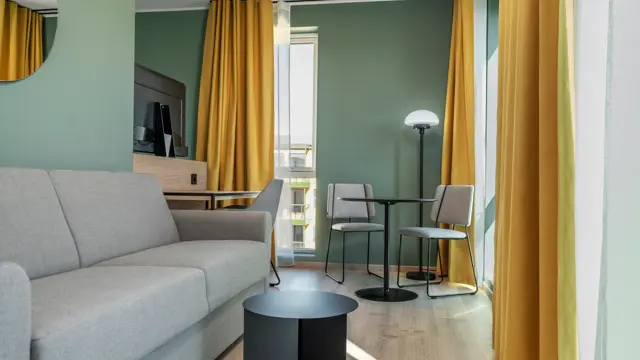 Standard queen rom med grå sofa, spiseplass, vinduer, gule gardiner og grønne vegger.