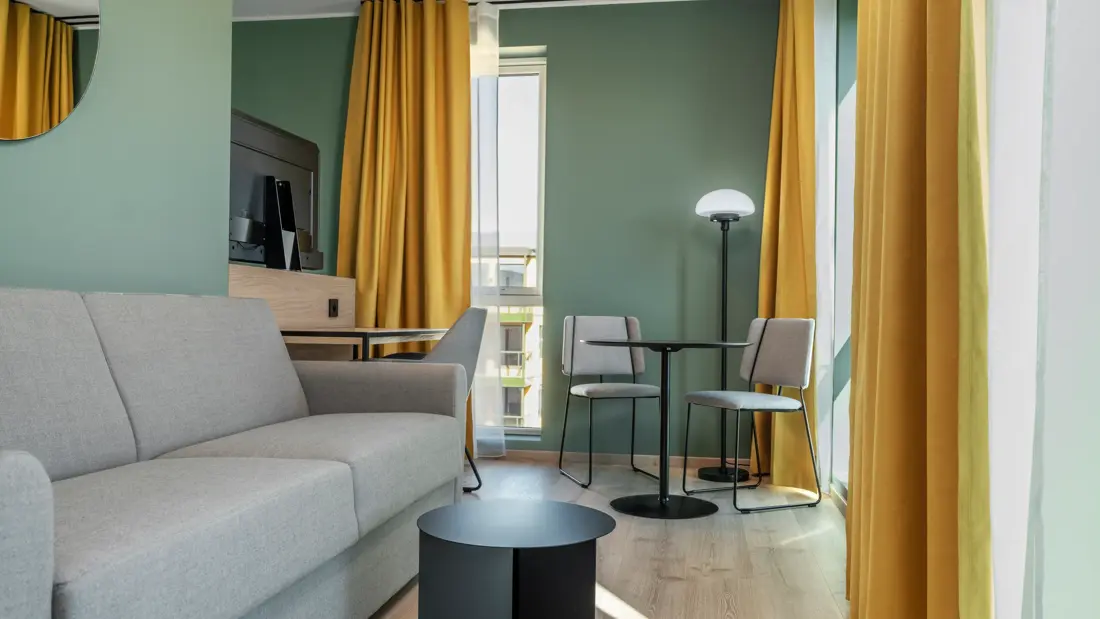 Standard queen rom med grå sofa, spiseplass, vinduer, gule gardiner og grønne vegger.