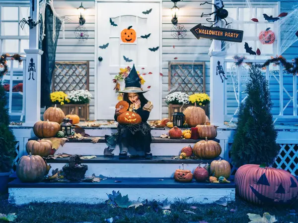 Little girl in witch costume sitting on the stairs in front of the house and holding Jack-o-Lantern Pumpkins on Halloween trick or treat