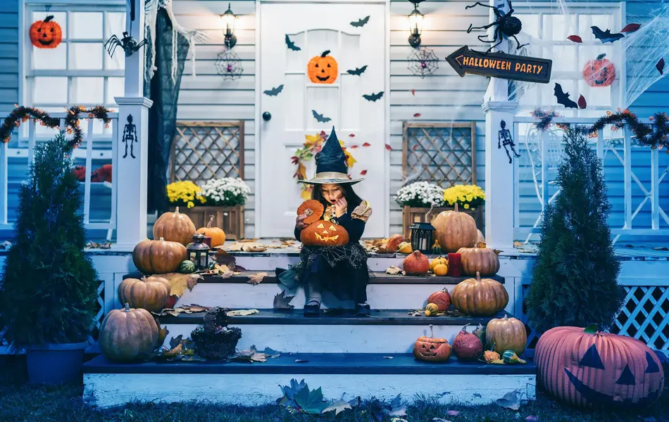 Little girl in witch costume sitting on the stairs in front of the house and holding Jack-o-Lantern Pumpkins on Halloween trick or treat