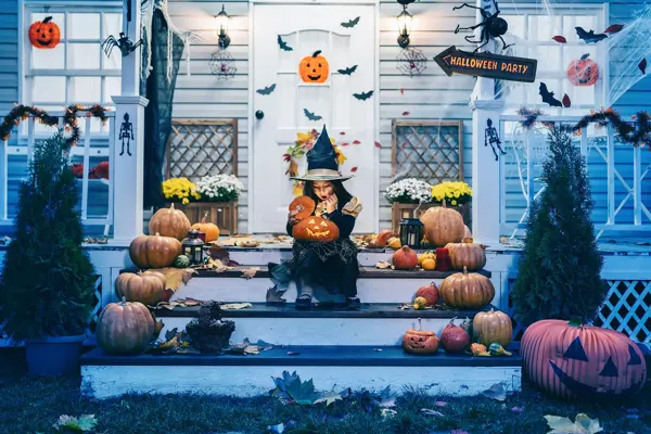 Little girl in witch costume sitting on the stairs in front of the house and holding Jack-o-Lantern Pumpkins on Halloween trick or treat
