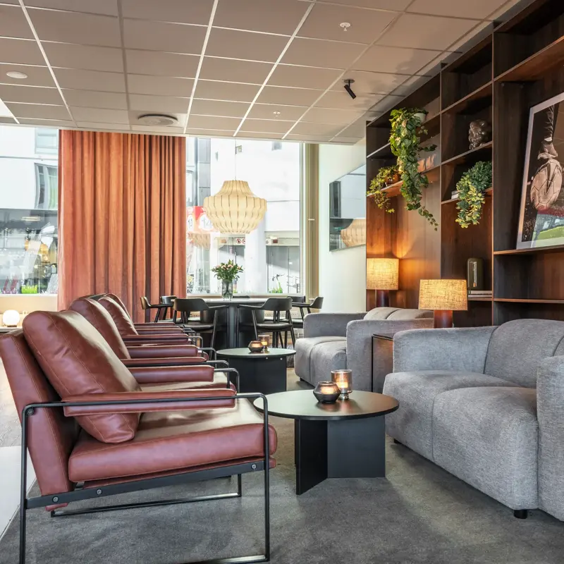 Lobbyområde med flere sittegrupper, grå sofaer og rød skinnstoler. Store vinduer og gardiner