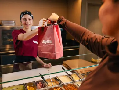 Ansatt overrekker en take away-pose til kunde ved en serveringsdisk.