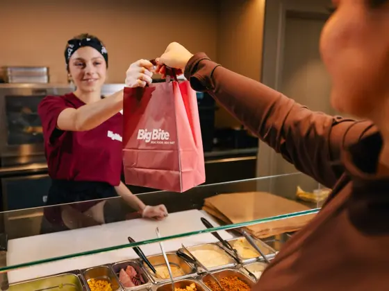 Ansatt overrekker en take away-pose til kunde ved en serveringsdisk.