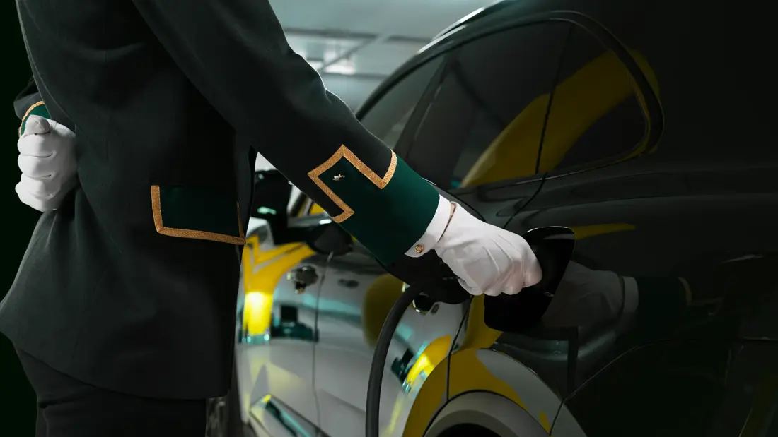 Ansatt i uniform og hvite hansker holder i en el-bil lader som står i en mørk bil