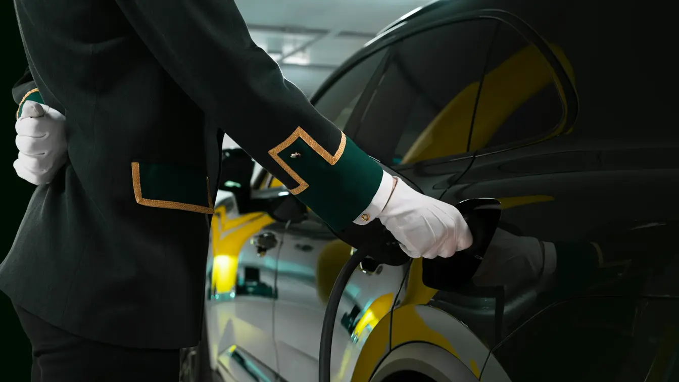 Ansatt i uniform og hvite hansker holder i en el-bil lader som står i en mørk bil