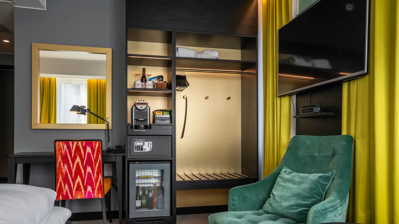 Detaljer på Superiorrom, skrivebord med oransje stol, stort speil, skap med minibar og Nespresso maskin, klesskap, vegghengt TV og en grønn lenestol.