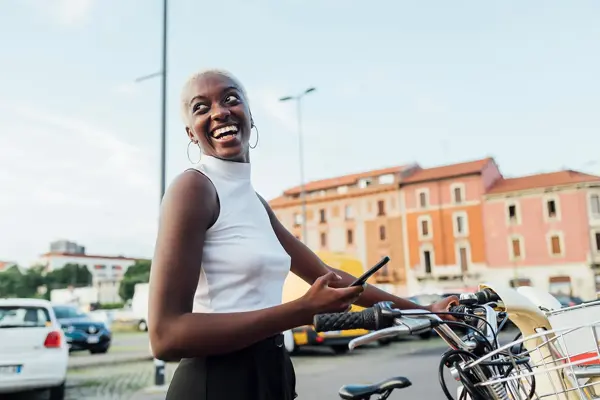 Halvportrett av dame med hvit tanktop som holder mobil og en sykkel med bybilde i bakgrunnen. 