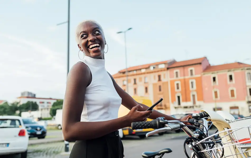 Halvportrett av dame med hvit tanktop som holder mobil og en sykkel med bybilde i bakgrunnen. 