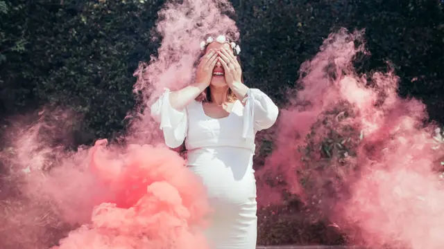 Happy pregnant woman covering her face with smoke grenade outdoors. Happy young expecting mother standing outside with smoke grenade.