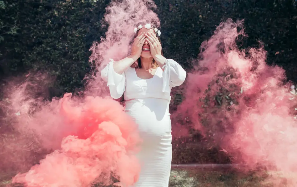 Happy pregnant woman covering her face with smoke grenade outdoors. Happy young expecting mother standing outside with smoke grenade.