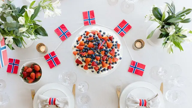 Fugleperspektiv ned på dekket bord, med hvit duk, fine tallerkener, kake og flagg i rødt, hvitt og blått, friske vårblomster. Foto til artikkel om borddekking på 17. mai.