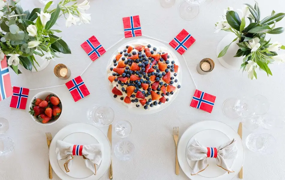 Fugleperspektiv ned på dekket bord, med hvit duk, fine tallerkener, kake og flagg i rødt, hvitt og blått, friske vårblomster. Foto til artikkel om borddekking på 17. mai.
