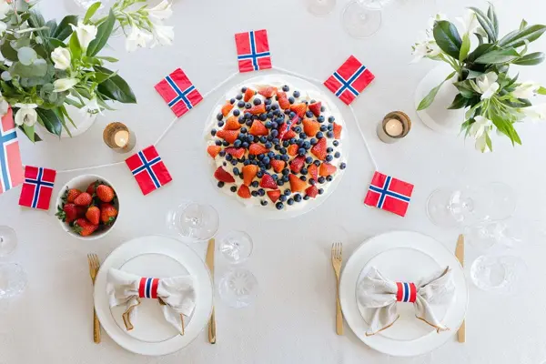 Fugleperspektiv ned på dekket bord, med hvit duk, fine tallerkener, kake og flagg i rødt, hvitt og blått, friske vårblomster. Foto til artikkel om borddekking på 17. mai.
