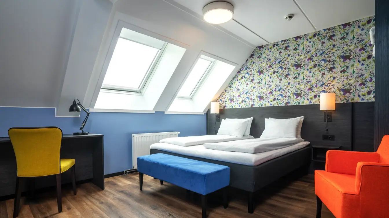 Blomstertapet, dobbeltseng, oransje stol, gul stol, parkettgulv, vindu.