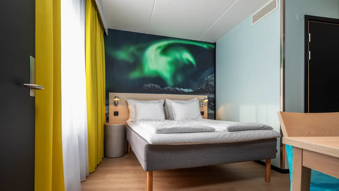 Dobbeltseng i junior suite med trehvit sengegavl, bilde av nordlys bak, lys grønn farge på veggen og gule gardiner.