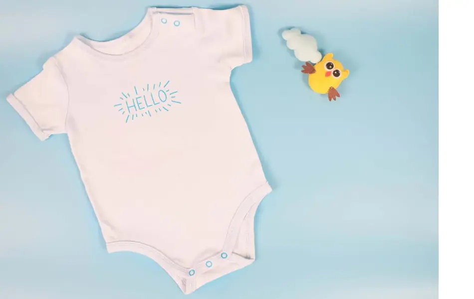 En hvit body til nyfødt som det står "Hello" på. Liten, gul leke ved siden av. Lyseblå bakgrunn. Illustrasjonsfoto til artikkel om kaker til babyshower.