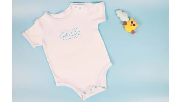 En hvit body til nyfødt som det står "Hello" på. Liten, gul leke ved siden av. Lyseblå bakgrunn. Illustrasjonsfoto til artikkel om kaker til babyshower.