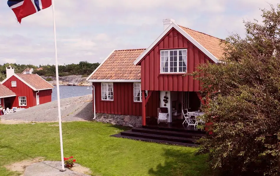 Oversiktsbilde over hage ved sjøen med familie som spiser ved et bord og flaggstang med norsk flagg. Foto til artikkel om 17. mai-dekorasjoner.