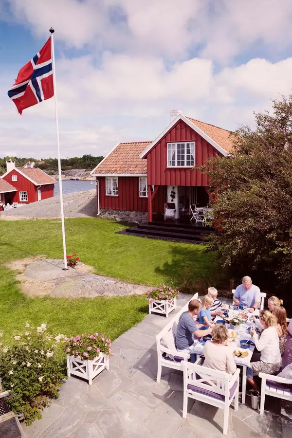 Oversiktsbilde over hage ved sjøen med familie som spiser ved et bord og flaggstang med norsk flagg. Foto til artikkel om 17. mai-dekorasjoner.