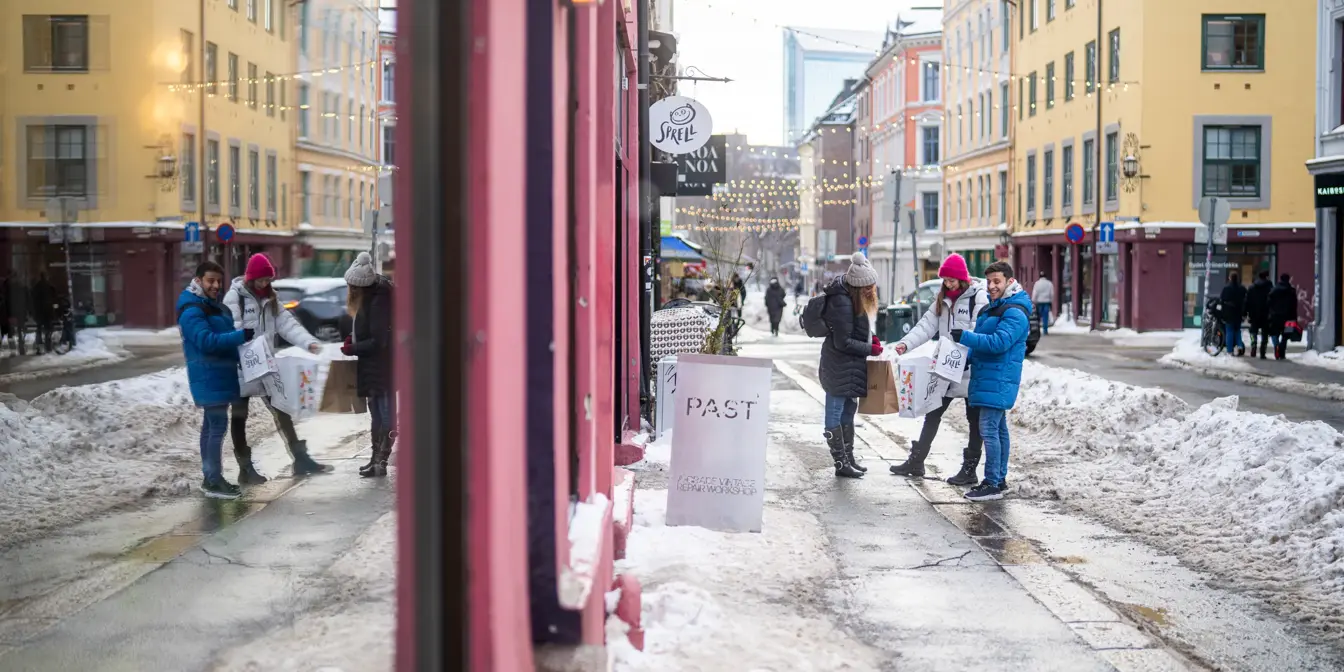 Tre venner med handleposer står på en snødekt gate med julelys og fargerike bygårder i Oslo, speilet i et butikkvindu.