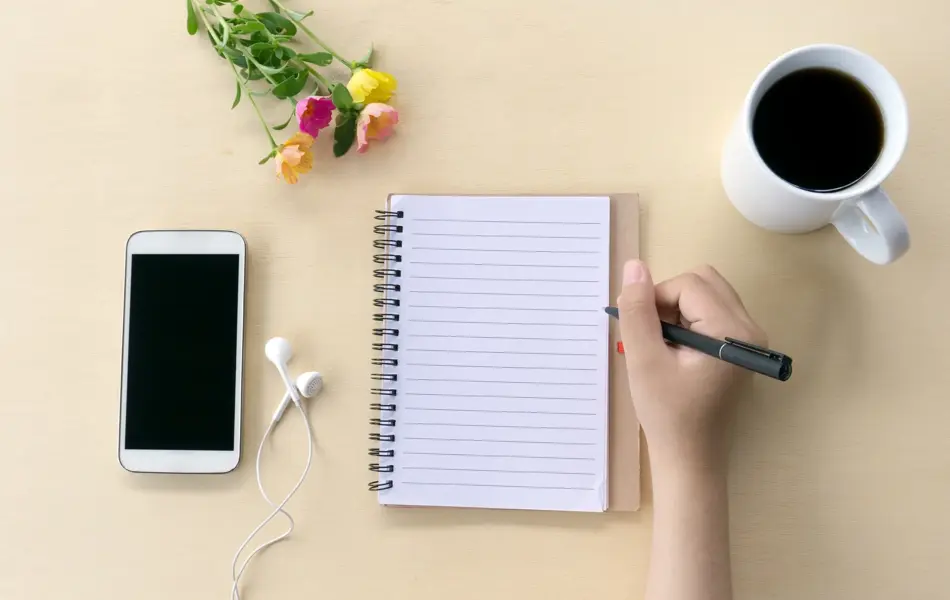 Directly above shot of an hands holding a pen on office desk with smart phone, notepad, flower, and cup of black coffee.