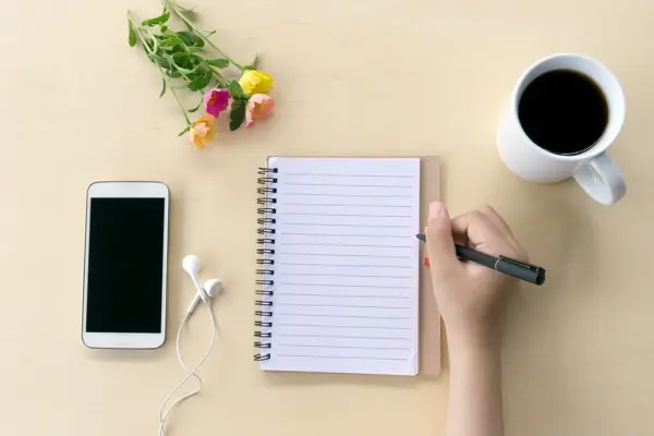 Directly above shot of an hands holding a pen on office desk with smart phone, notepad, flower, and cup of black coffee.