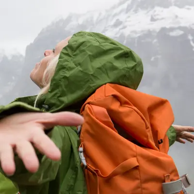 Dame i grønnregnjakke med oransje sekk holder armene ut og ser opp mot himmelen med snødekkede fjell i bakgrunnen